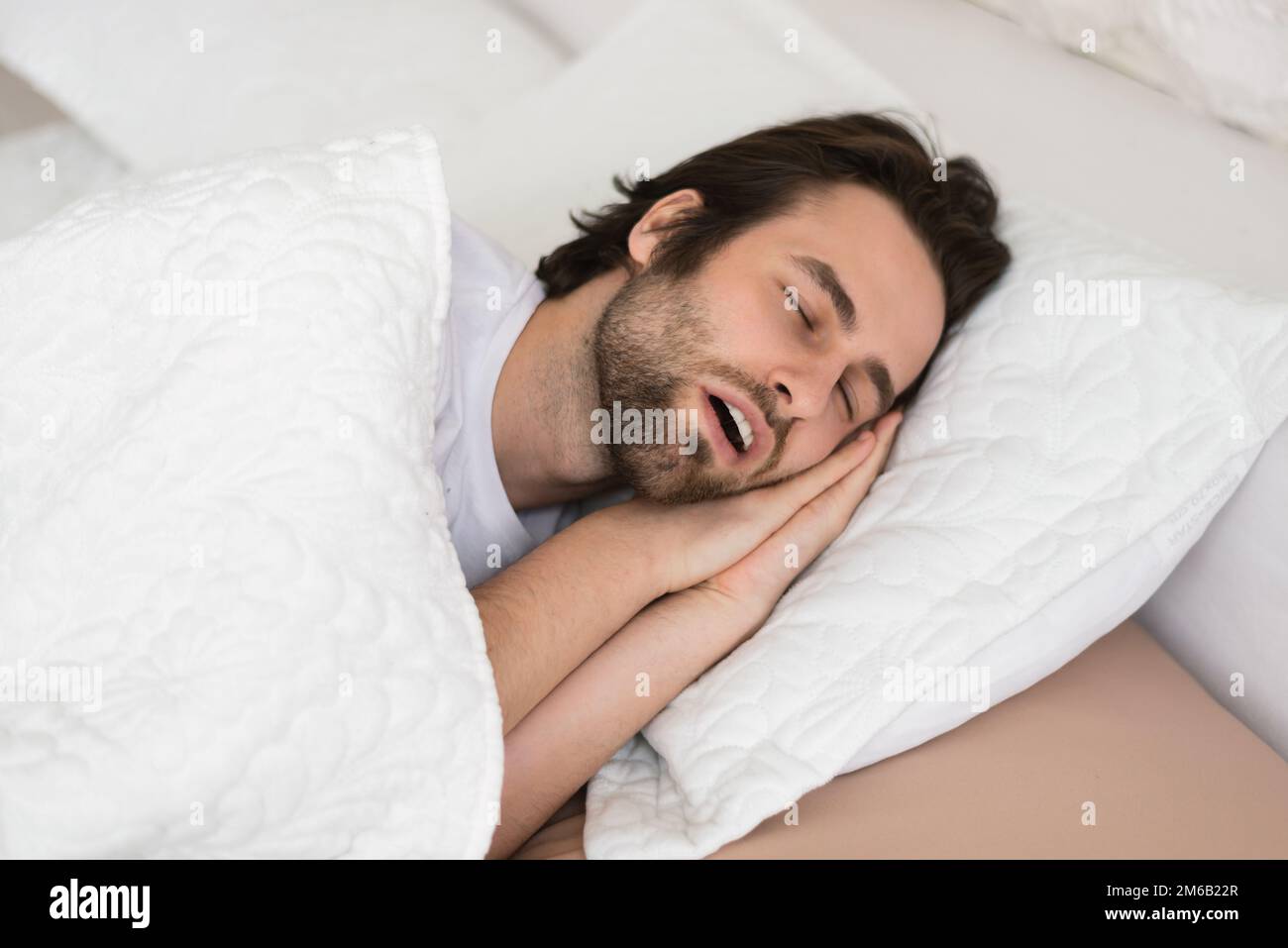 Tired young caucasian male with open mouth sleeping on white soft bed in bedroom in spare time