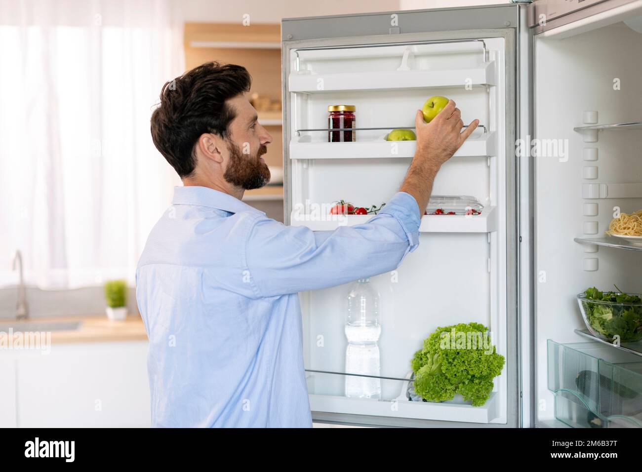 Happy hungry millennial caucasian bearded guy opens refrigerator door and takes green apple for cooking food Stock Photo