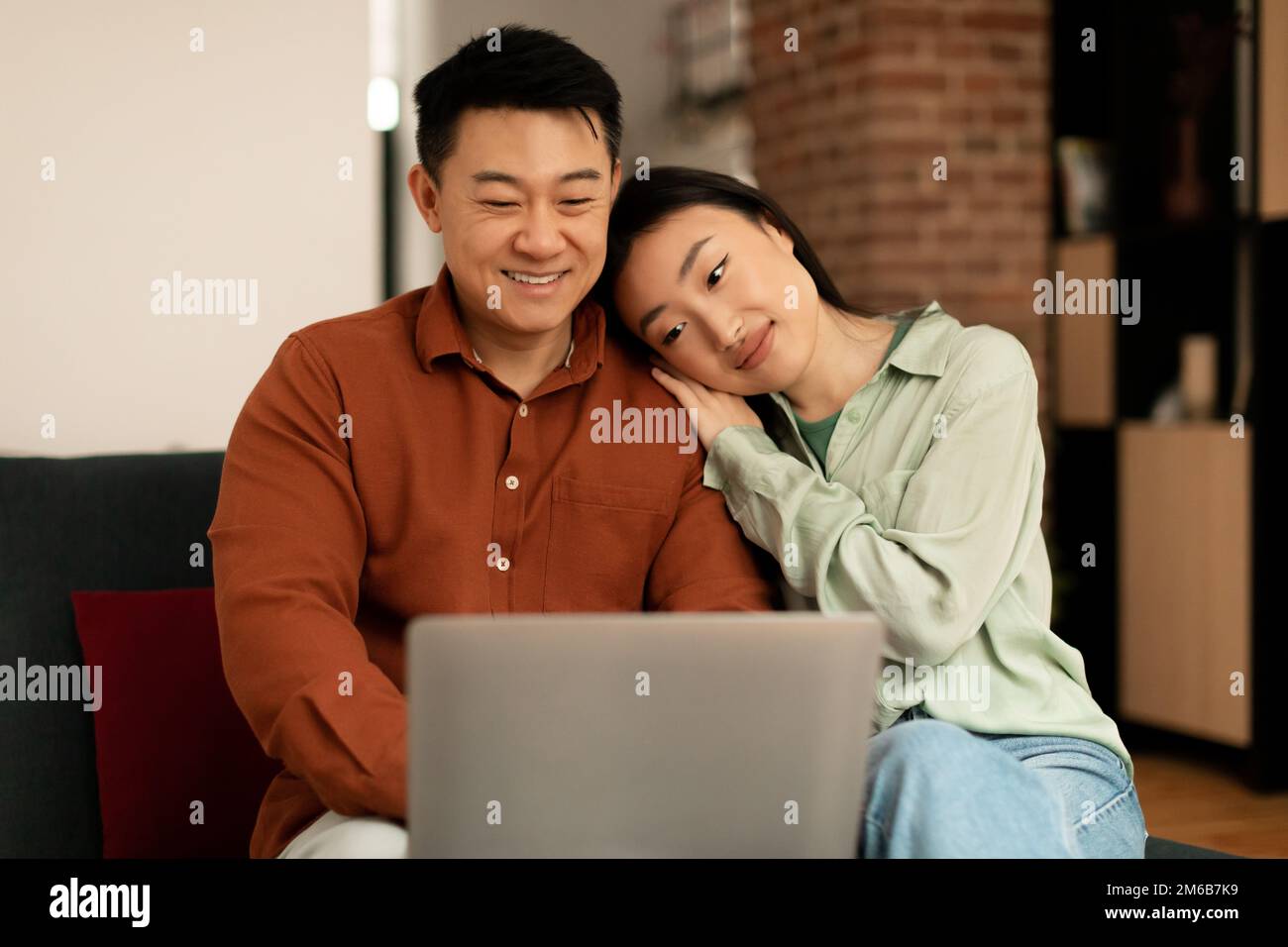 Loving asian couple using laptop together, surfing internet or watching movie online, wife leaning on husband's shoulder Stock Photo