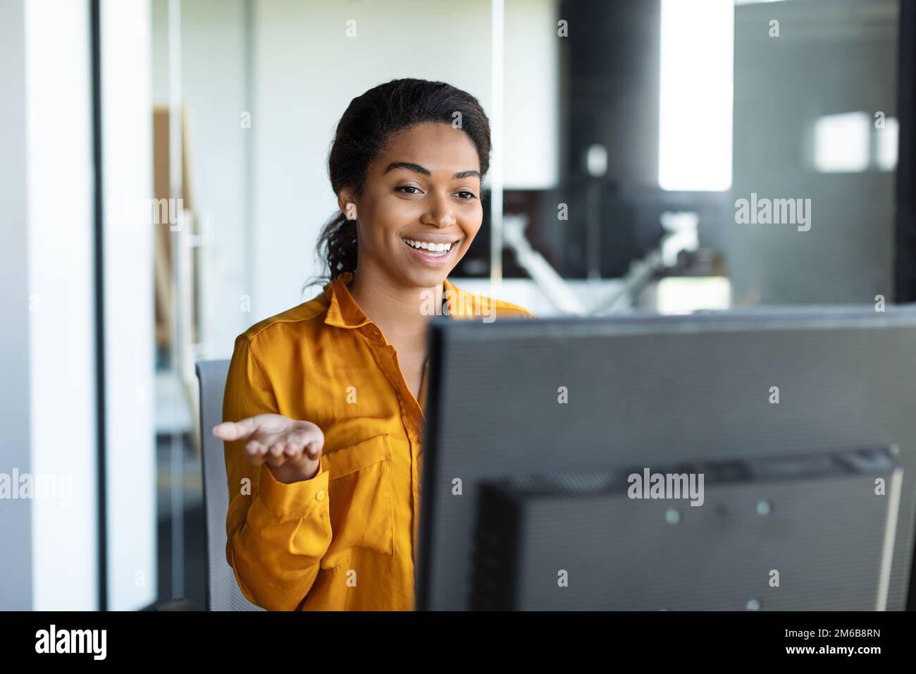 Positive black female entrepreneur gesticulating at computer webcamera, working and having video call in office Stock Photo