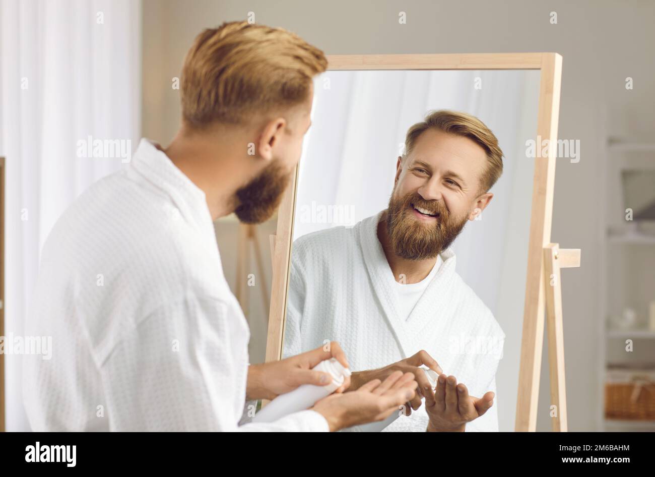 Happy bearded man with beard lotion in his hands enjoys amorning routine of self-care. Stock Photo