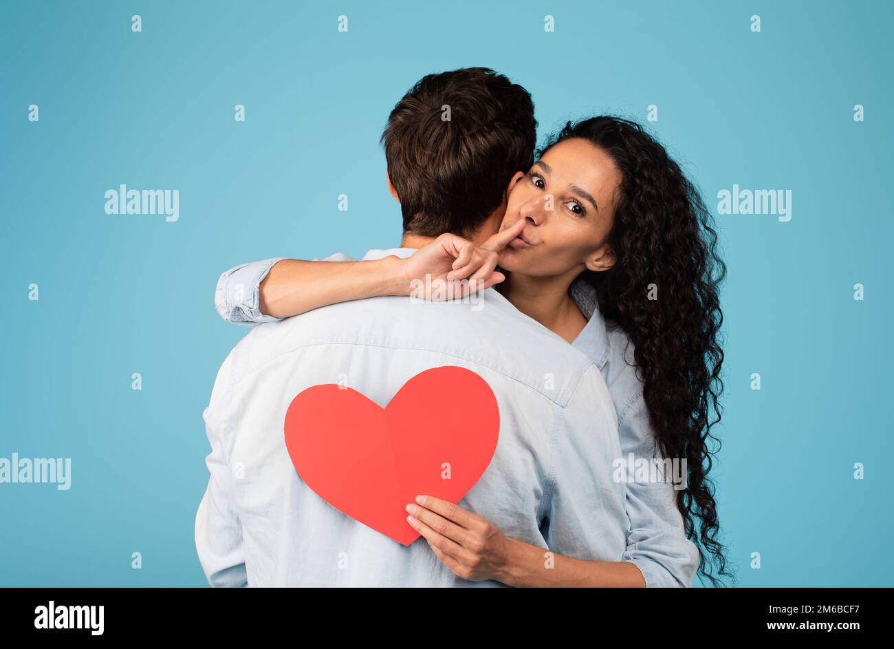Cheerful young arab wife hugs european husband, holds heart, shows finger sign shhh on blue background Stock Photo