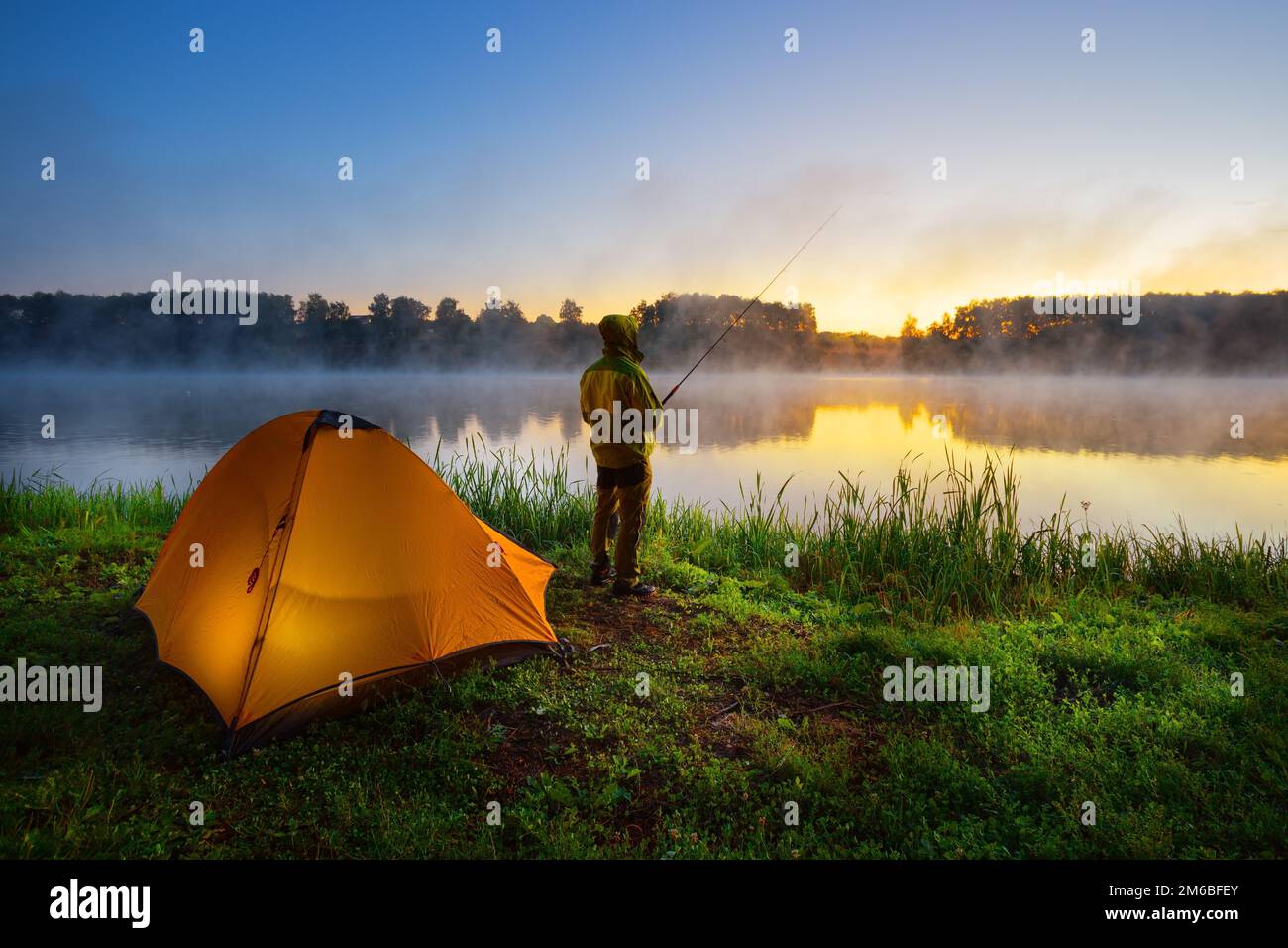 Fisherman on the bank of foggy river near an orange tent in the early morning Stock Photo