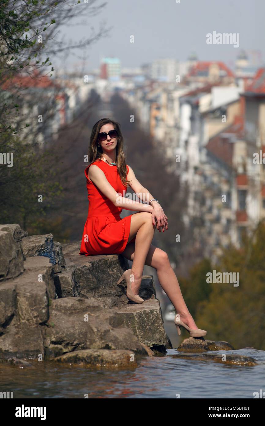 Beautiful blonde woman with orange dress sitting on a rock in a park ...