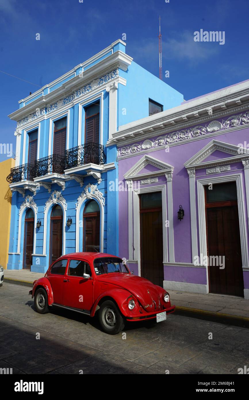 A bright red Volkswagen Beetle drives past restored purple and blue heritage houses on Calle 59 a famous street in Merida Centro, Yucatan, Mexico Stock Photo