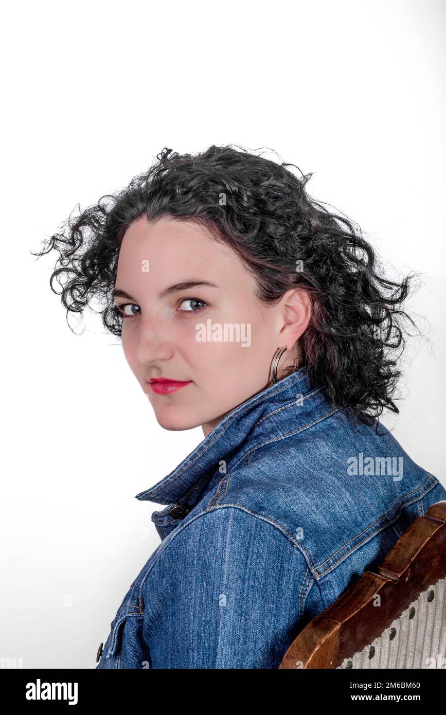 Young model in her mid twenties sitting on a wooden chair with a blue denim jacket. Stock Photo