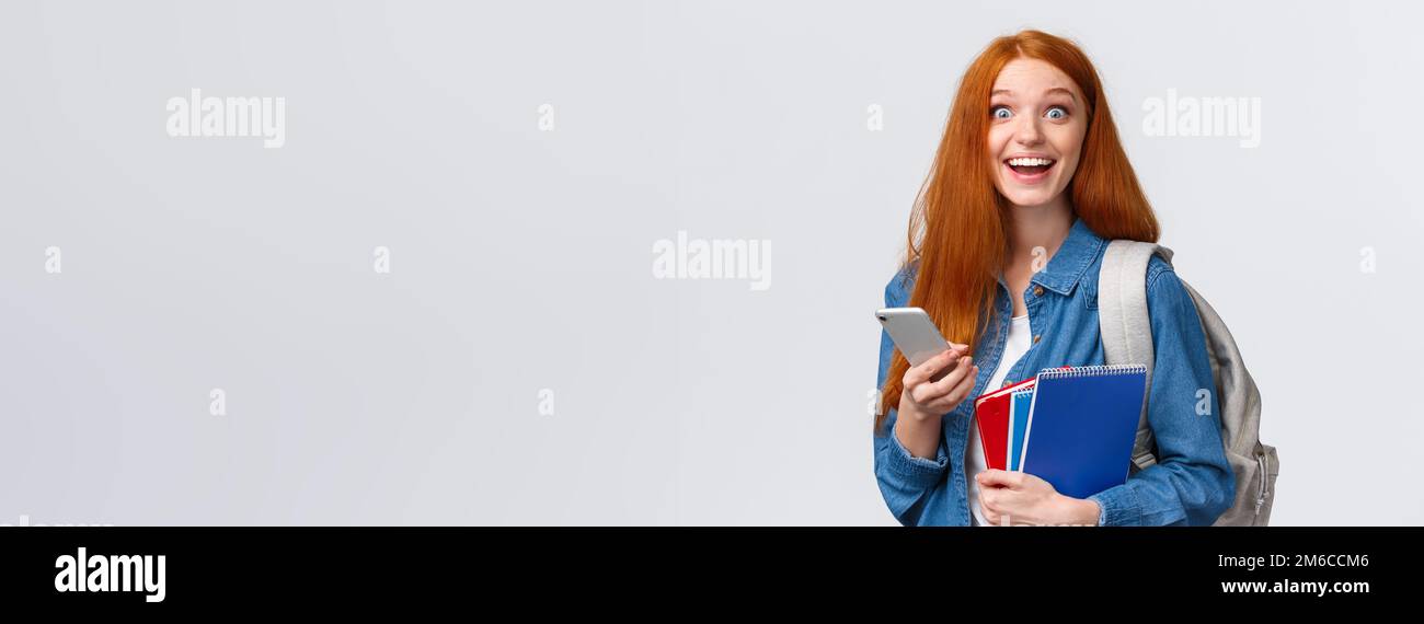 Excited and happy, relieved cute redhead female college student with backpack, notes in notebook and smartphone, looking cheerful camera, texting Stock Photo
