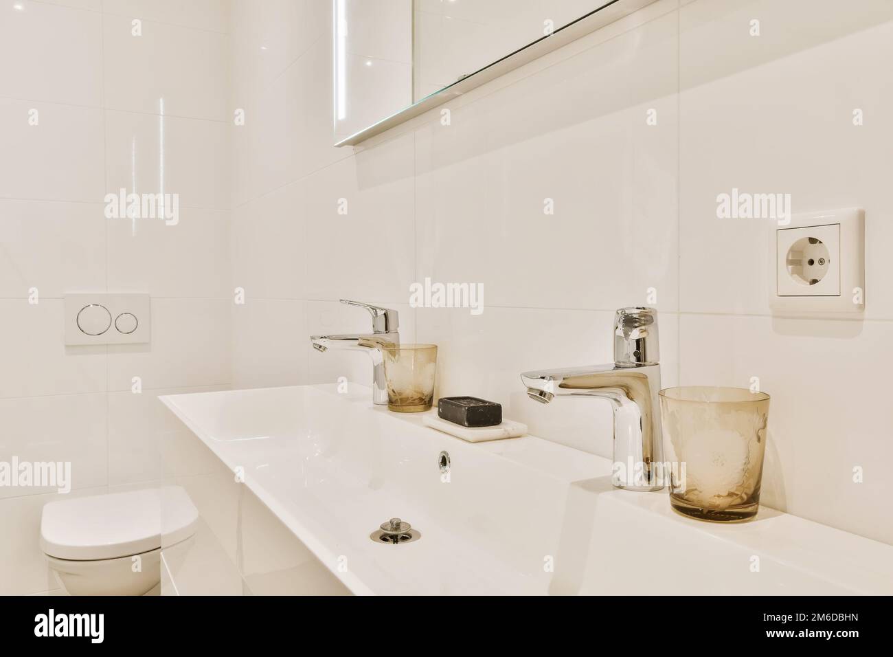 a modern bathroom with white tile walls and flooring, including the sink and toilet in the room is illuminated by a large mirror Stock Photo
