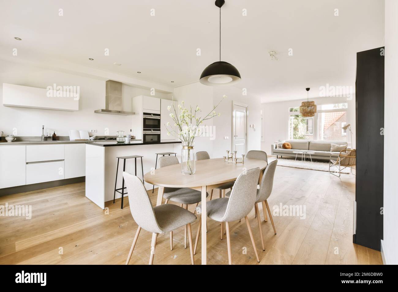 a kitchen, dining room and living area in an apartment with white walls and wood flooring the table is surrounded by grey chairs Stock Photo
