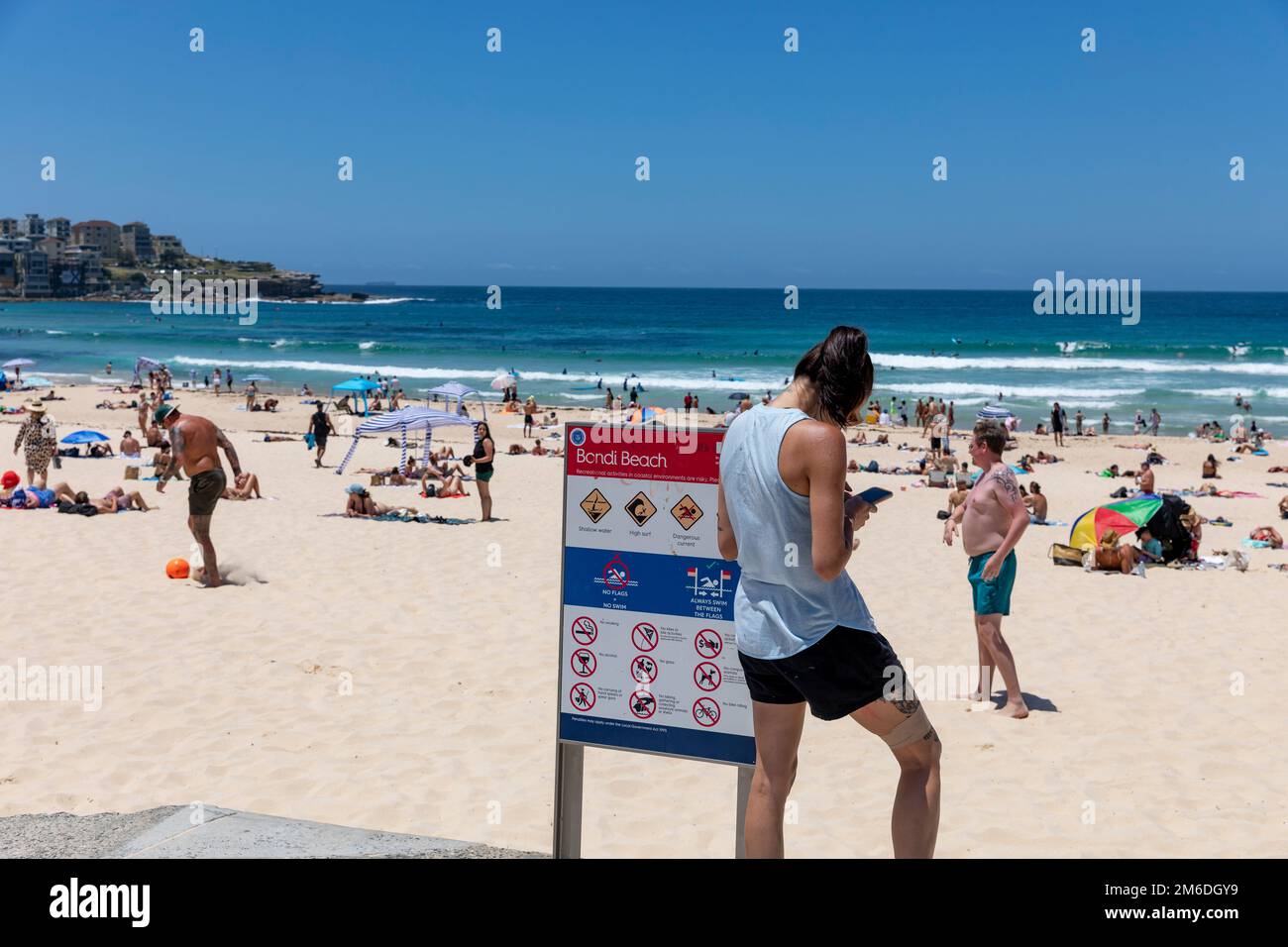 Bondi Beach Sydney 2023, young brunette woman texting sms on her mobile phone, leg and arm tattoos, stood beside Bondi Beach sign,Sydney,NSW,Australia Stock Photo