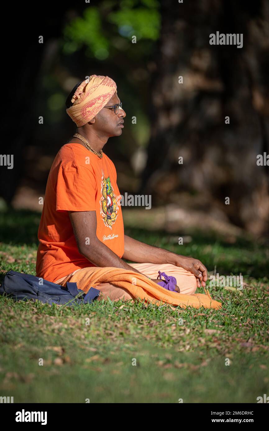 A young man, with head-dress, sits meditiating, cross-legged on a grassy field in the afternoon sun in Macintosh Park, Surfers Paradise, Australia. Stock Photo
