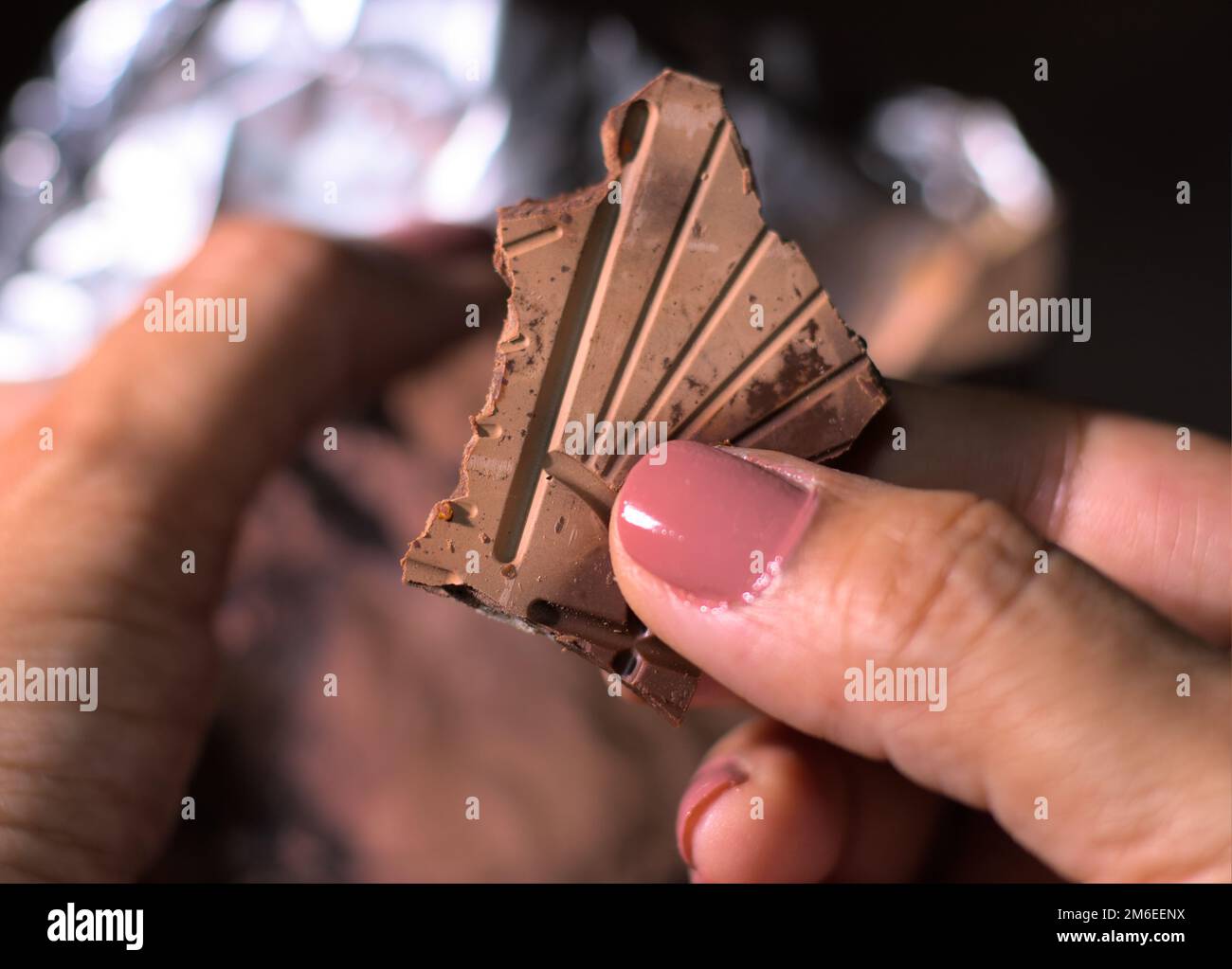 Chunk or piece of a dark chocolate in a female hand. Out of focus Silver foil in background. natural candid photography. Night. Stock Photo