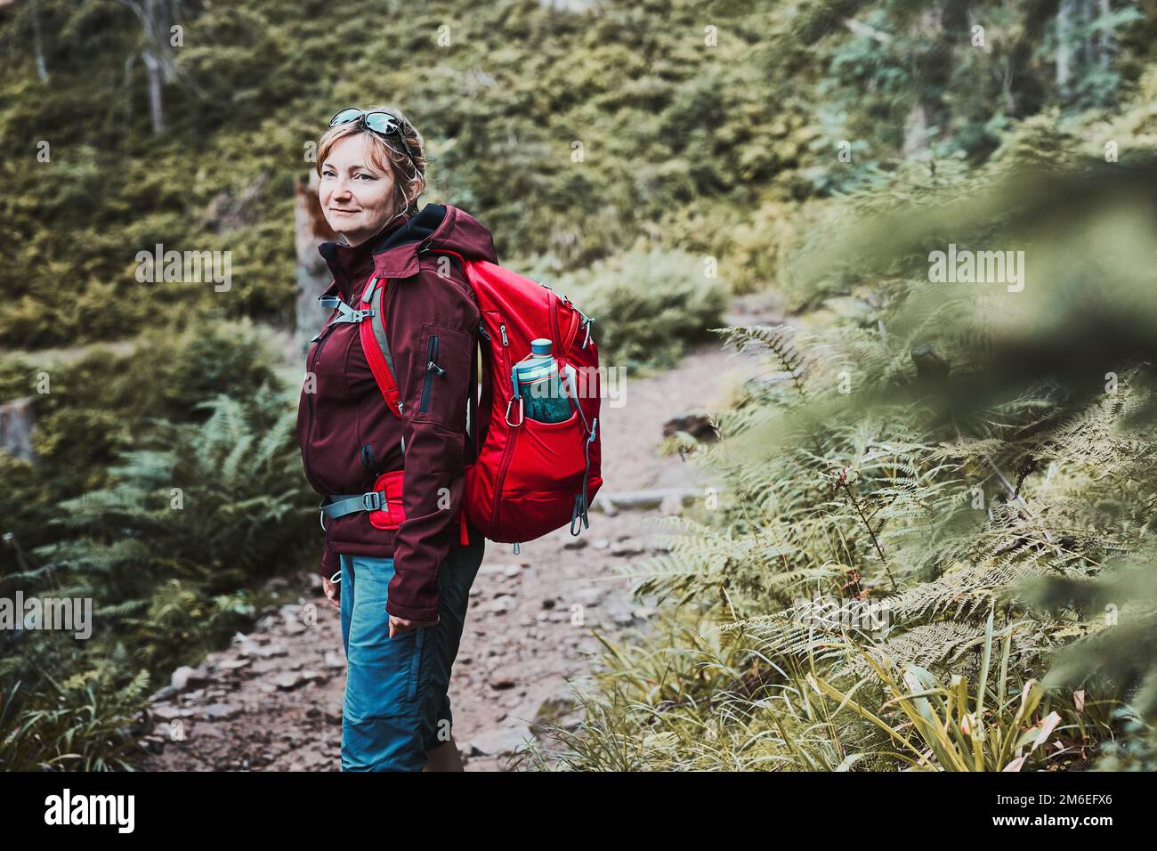 Woman with backpack hiking in mountains, spending summer vacation close to nature. Woman walking on path among bushes and trees Stock Photo