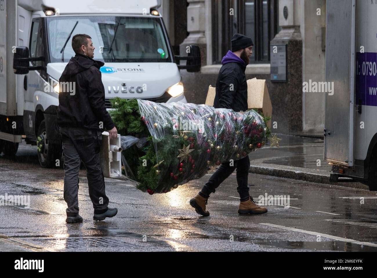 Removals workers carry a wrapped Christmas tree after retrieving it from an office building on the first day back in the New Year, City of London, UK Stock Photo
