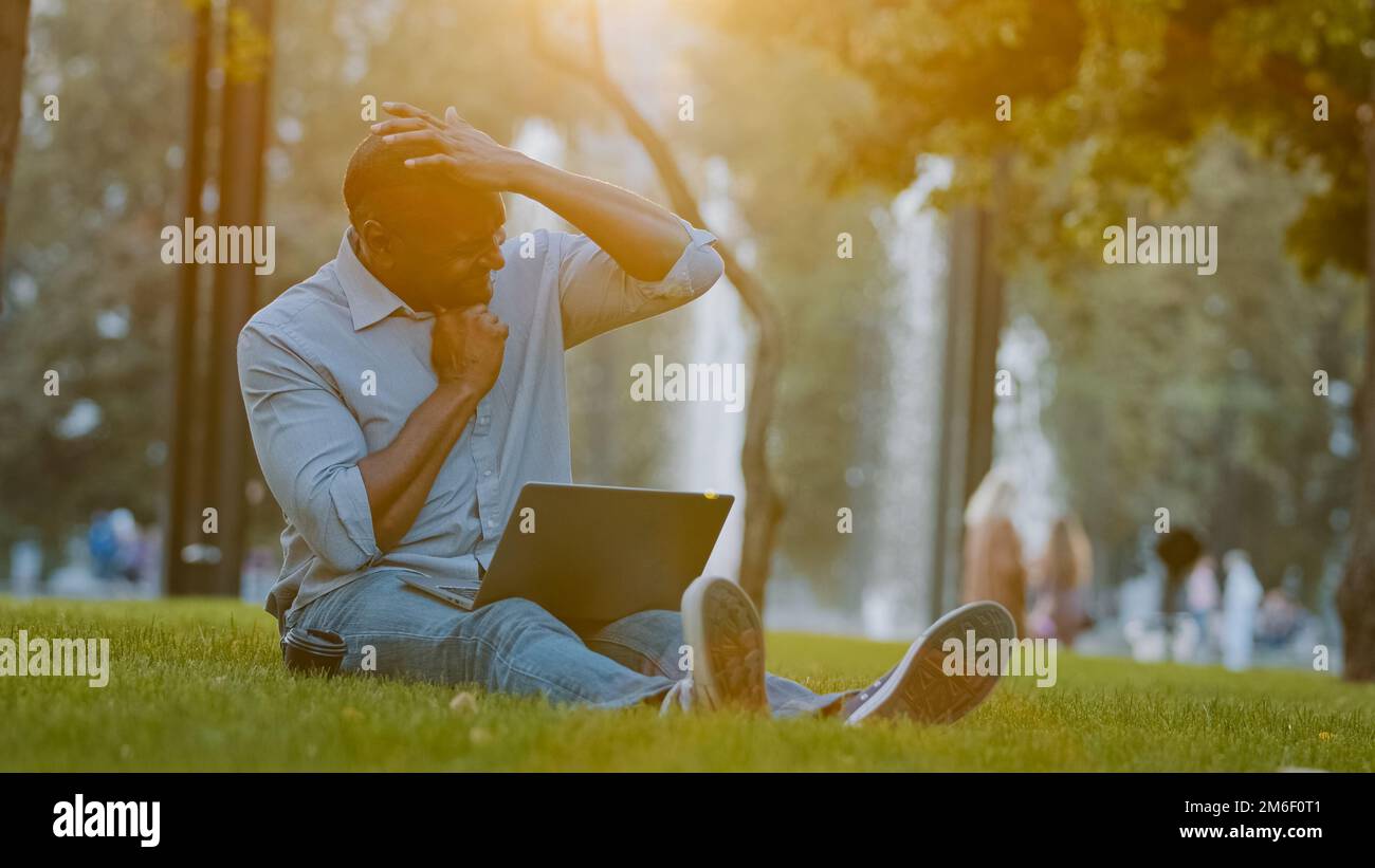 Elderly middle-aged African American businessman entrepreneur man sitting on grass in city park working on computer laptop feeling tired stressed Stock Photo
