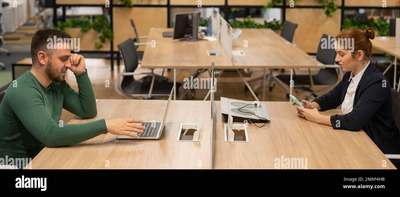 Red-haired caucasian woman uses a smartphone and a bearded man works behind a laptop in an open space office.  Stock Photo