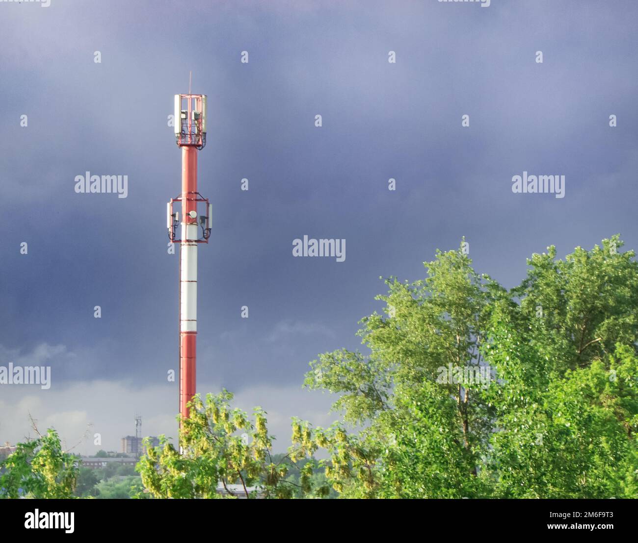 High tower of the GSM transmitter station in the evening against the background of blue sky and trees, in summer, after rain Stock Photo