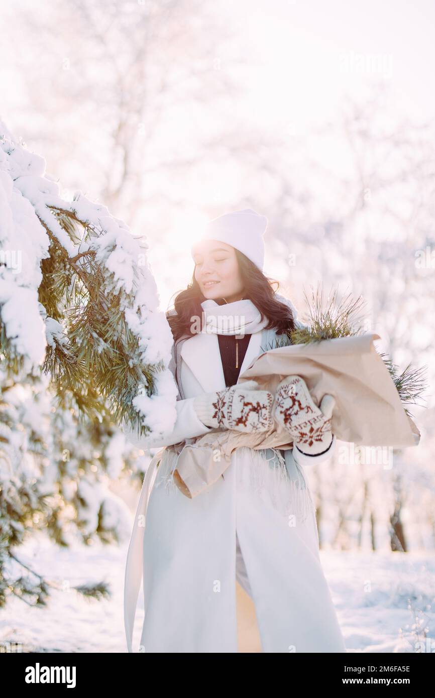 Happy young smiling woman walks in forest among snow covered pine trees in sunny winter day with bouquet in her hands. Stock Photo