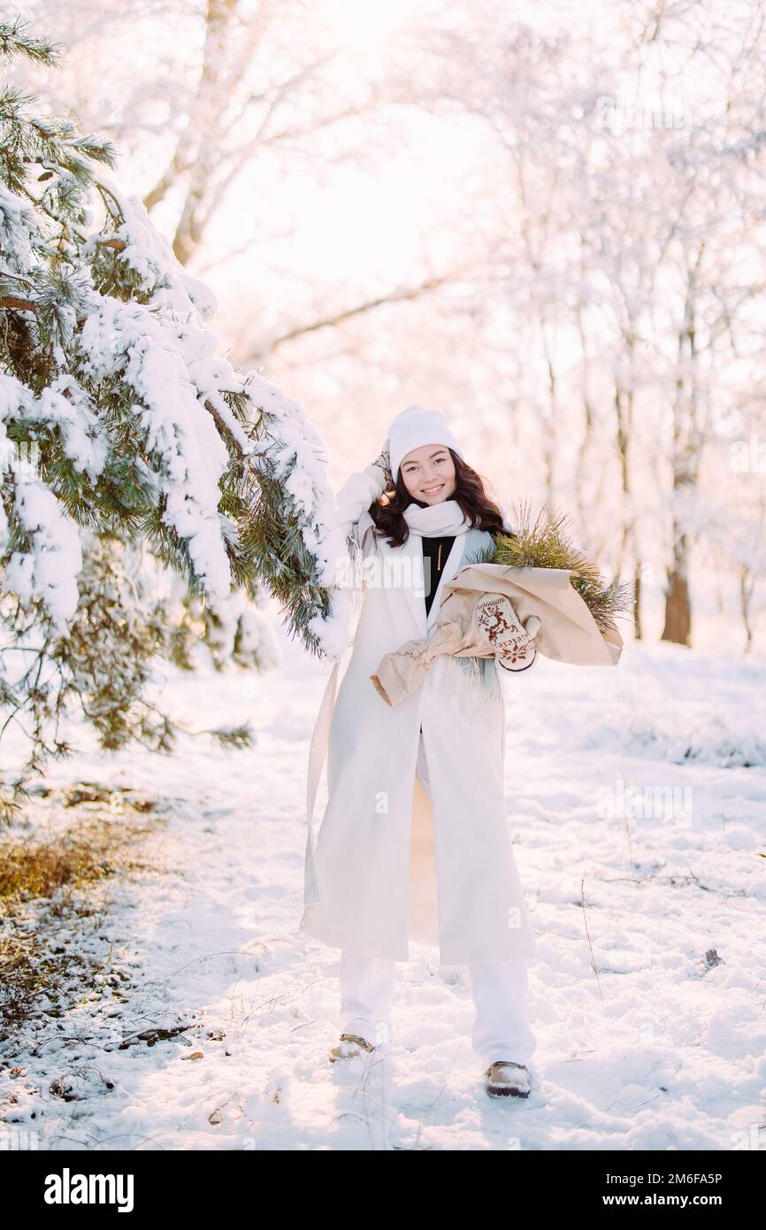 Happy young smiling woman walks in forest among snow covered pine trees in sunny winter day with bouquet in her hands. Stock Photo