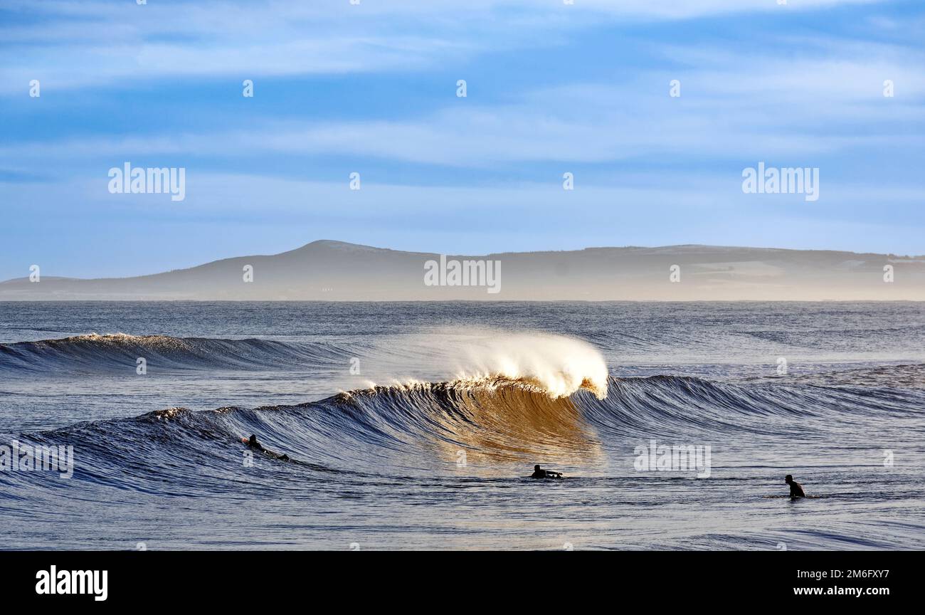 Lossiemouth East Beach Moray coast Scotland curling waves with spray and three surfers waiting for a larger wave Stock Photo