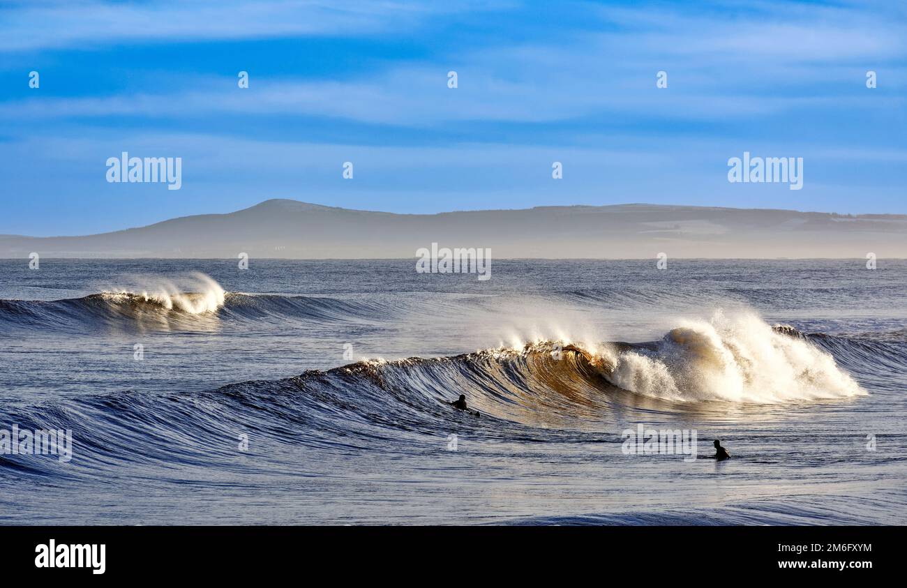 Lossiemouth East Beach Moray coast Scotland curling waves with spray and two surfers waiting for a larger wave Stock Photo