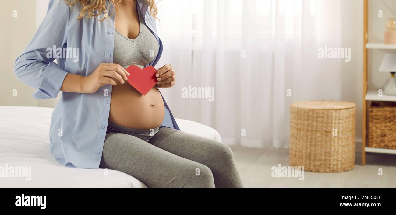 Loving expectant mother sitting on the bed and holding a paper heart in her hands Stock Photo
