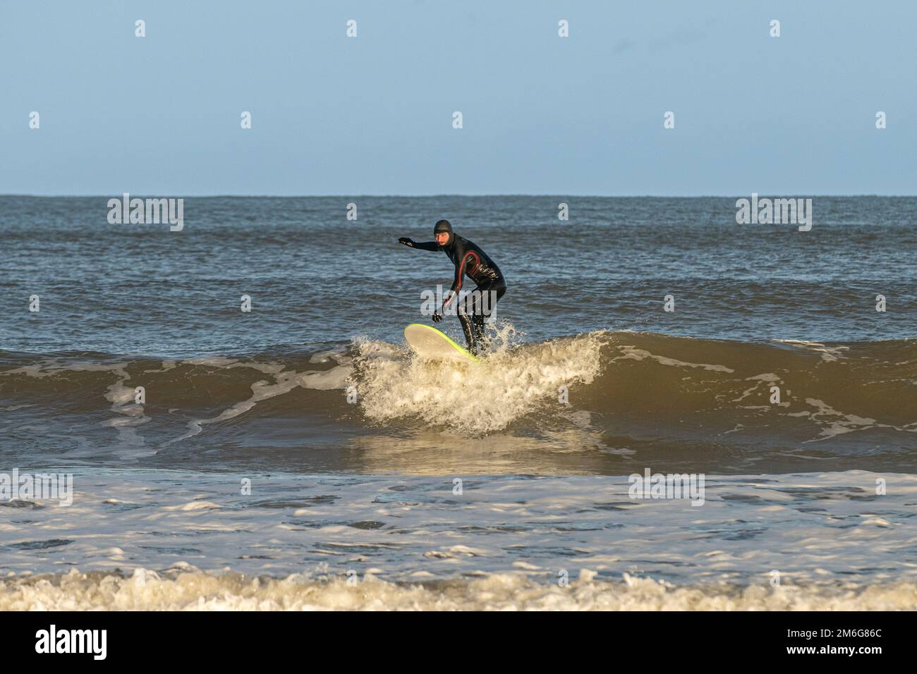 Caucasian male surfer wear a black wetsuit coming onto shore. Cayton Bay, North Yorkshire, UK Stock Photo