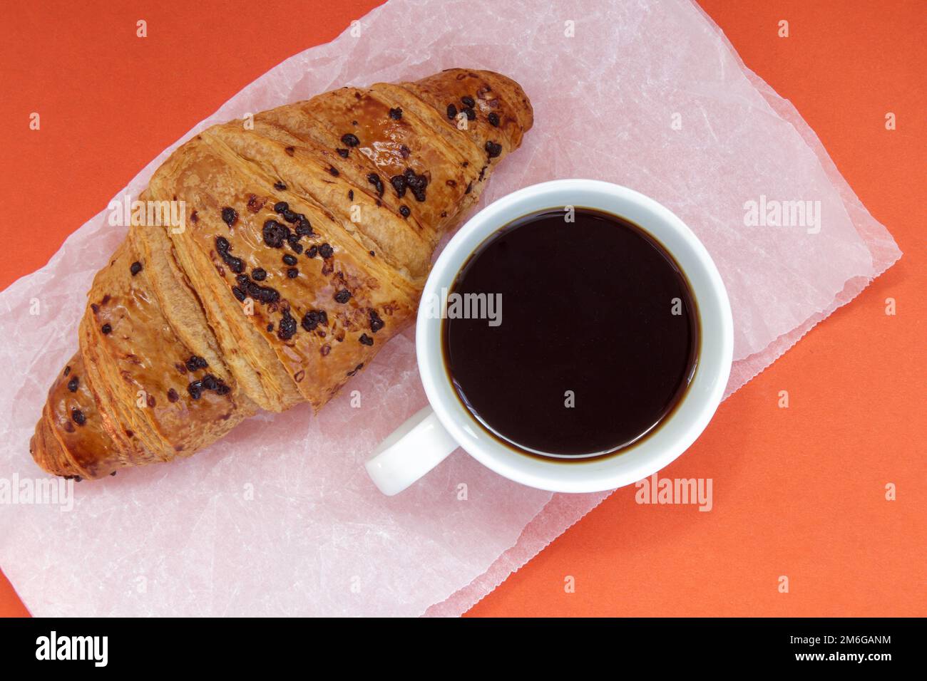 Black coffee without milk in a white cup and a chocolate croissant on parchment and bright background. French breakfast with fre Stock Photo