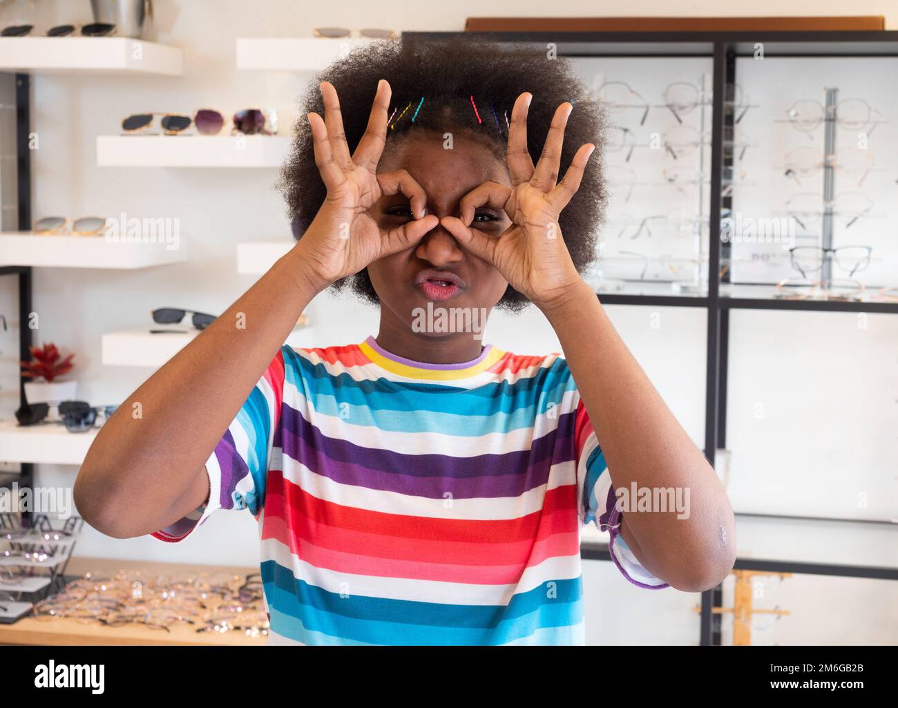 Portrait of a brightly smiling African girl puts her finger in a semicircle symbolizing eyeglasses as she selects a frame in an optical shop. Stock Photo