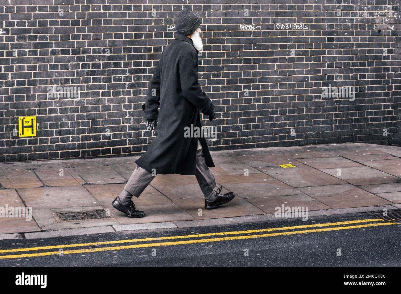A stylish old, bearded man with a black coat walking briskly in front of a grungy black wall with yellow lines on the street. London, UK Stock Photo