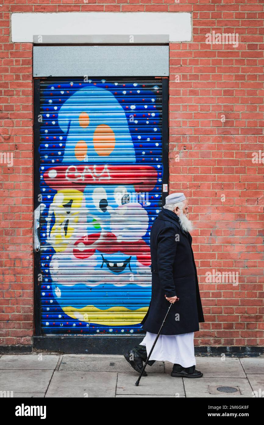 A bearded man with a cane and black coat walking in front of a colorful graffiti of a bearded head on a shutter in a red brick wall. London, UK Stock Photo