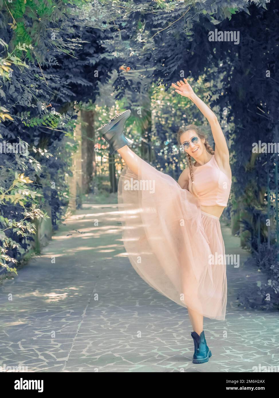 A lady with pigtails in a tutu and rough shoes makes a forward leg swing in an alley in a summer park. Stock Photo