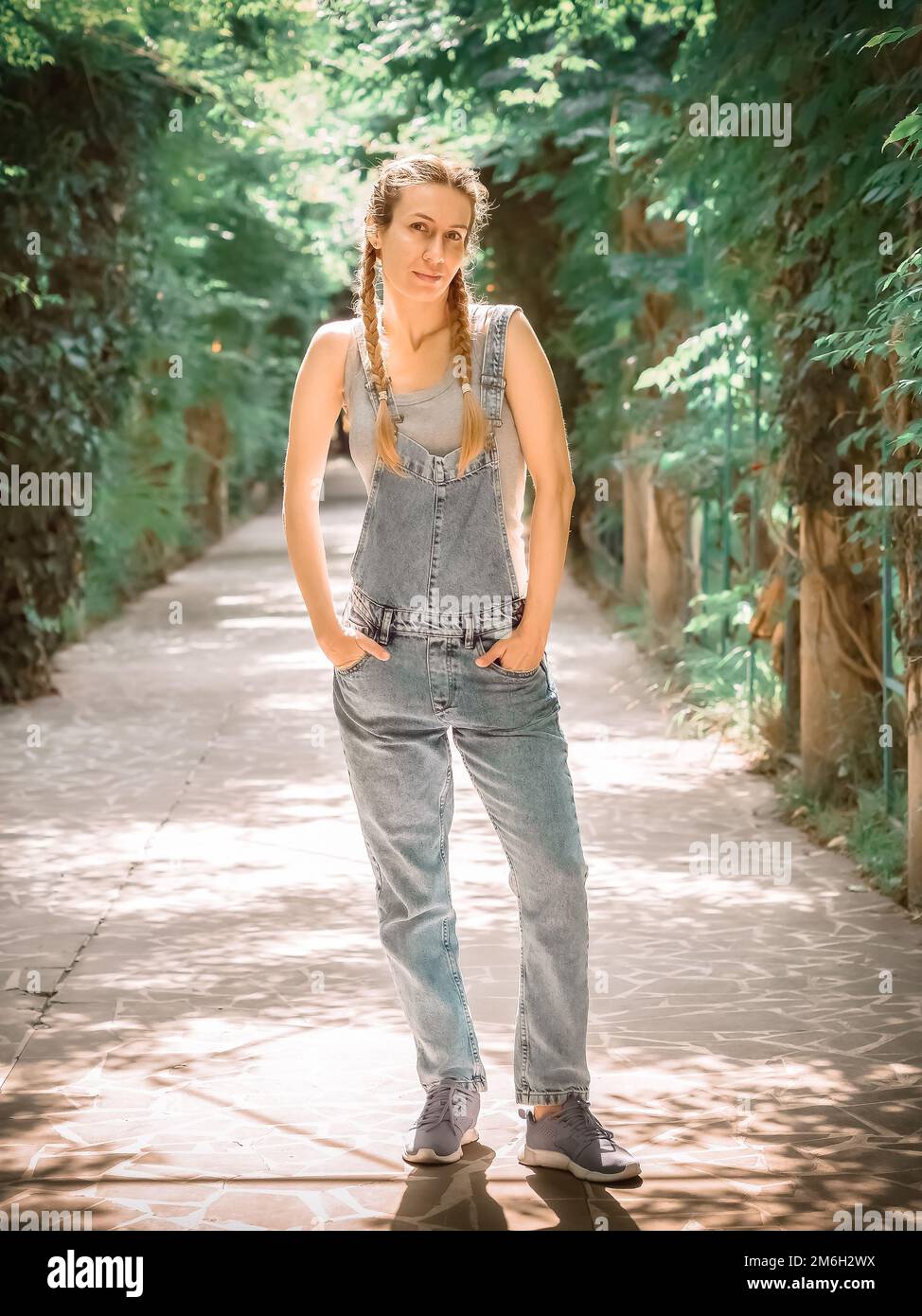 A girl with pigtails in a denim overalls stands with her hands in her pockets on a sunlit alley in the park Stock Photo
