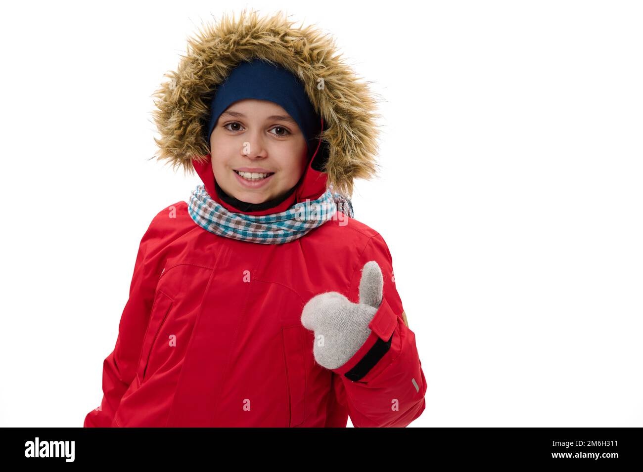 Cute teen boy in red down parka with fur hood, thumbing up, smiling, looking at camera, isolated over white background Stock Photo