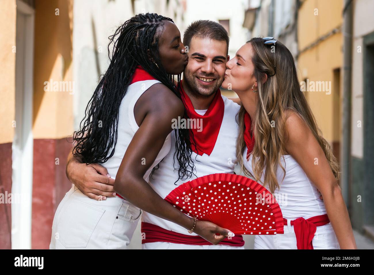 two friends of different ethnicity kiss their friend in the street during the popular festivities of a town in spain. concept of fun, friendship Stock Photo