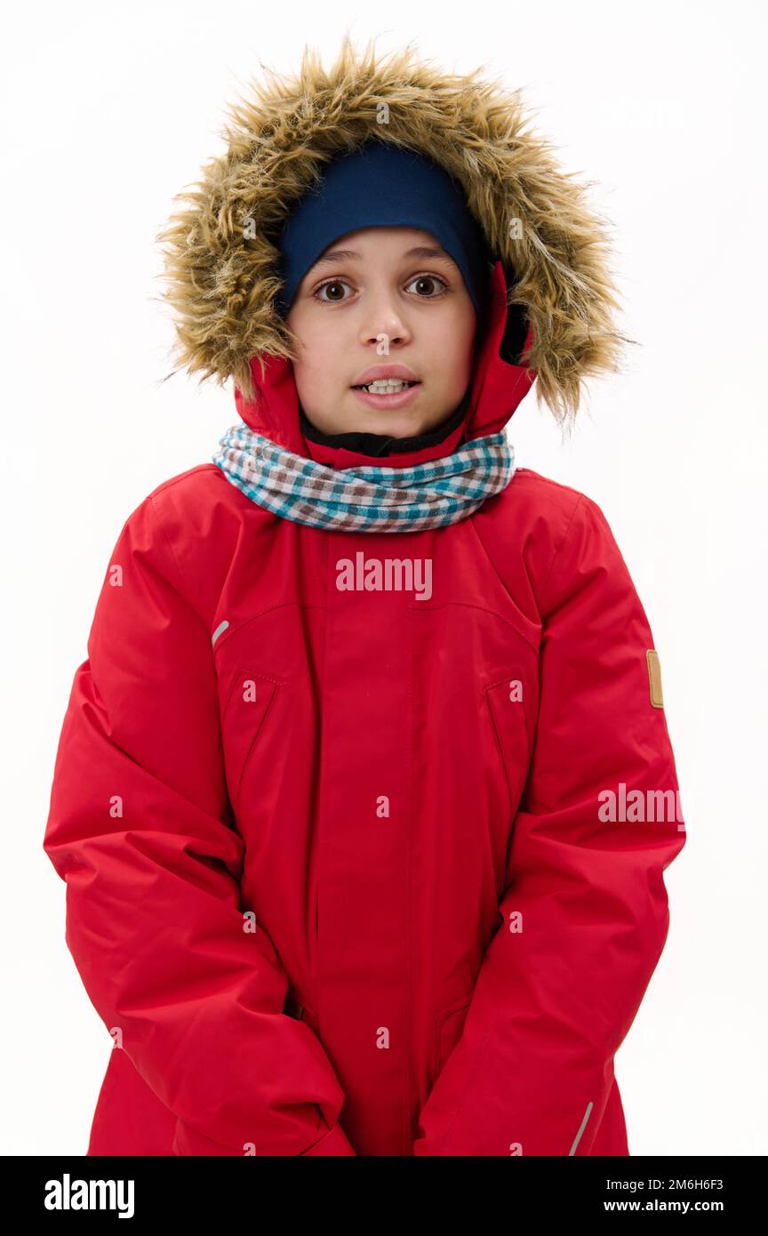 Vertical studio shot of a Middle-Eastern teenage boy, wearing red parka with fur hood and blue scarf, smiling at camera Stock Photo