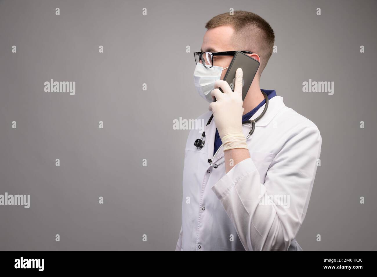 A young Caucasian surgeon in glasses talks on the phone, consults in a white surgical gown and a mask on a light background. Med Stock Photo