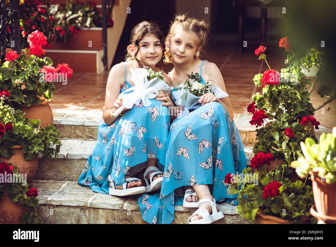 Two girls, sisters sitting outdoors in butterfly patterned dress, surrounded with flowers Stock Photo