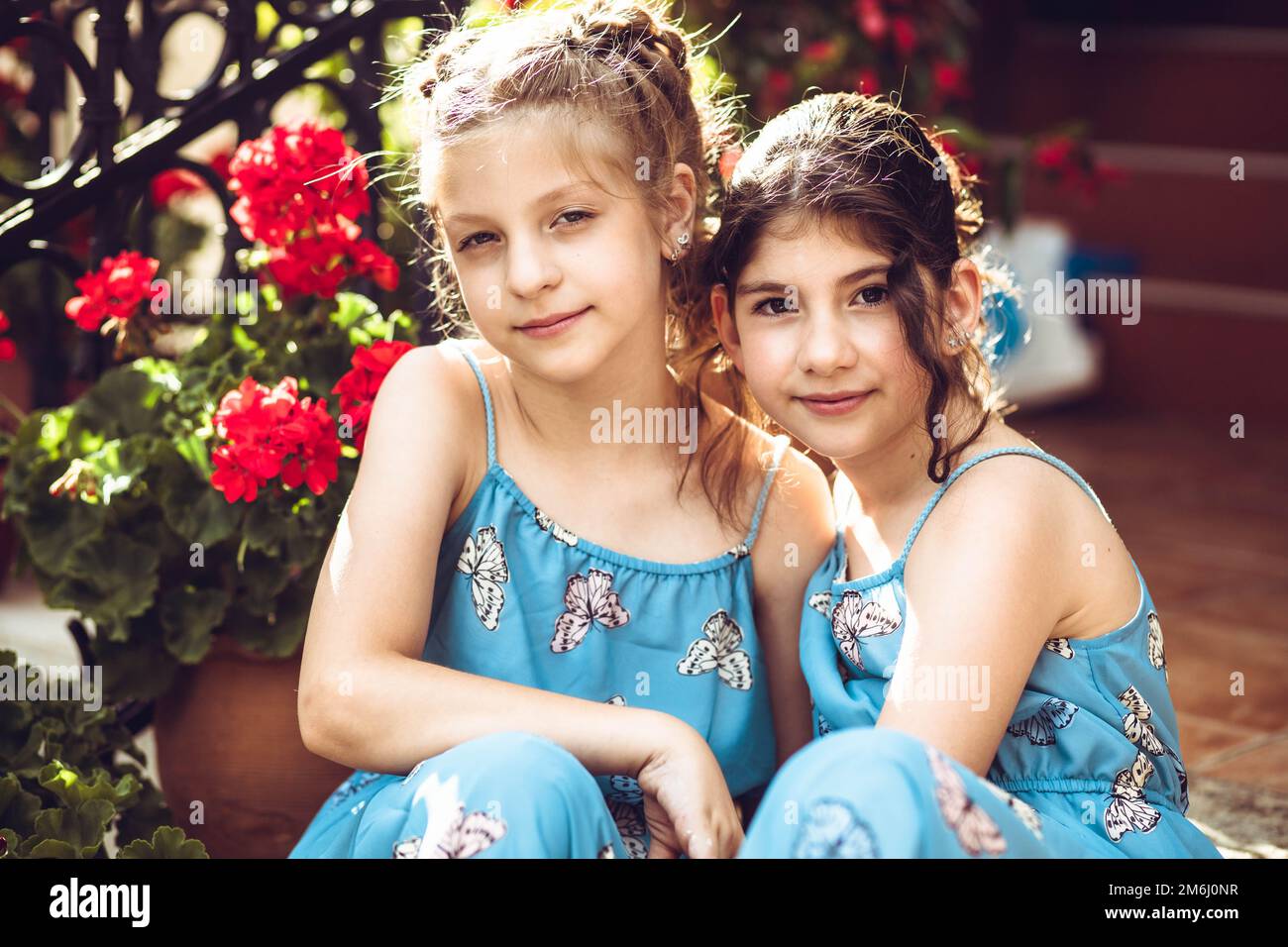 Two girls, sisters sitting outdoors in butterfly patterned dress, surrounded with flowers Stock Photo