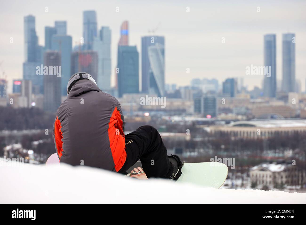 Snowboarder fastens binding of snowboard while sitting on background of skyscrapers of Moscow city. Leisure in winter park Stock Photo