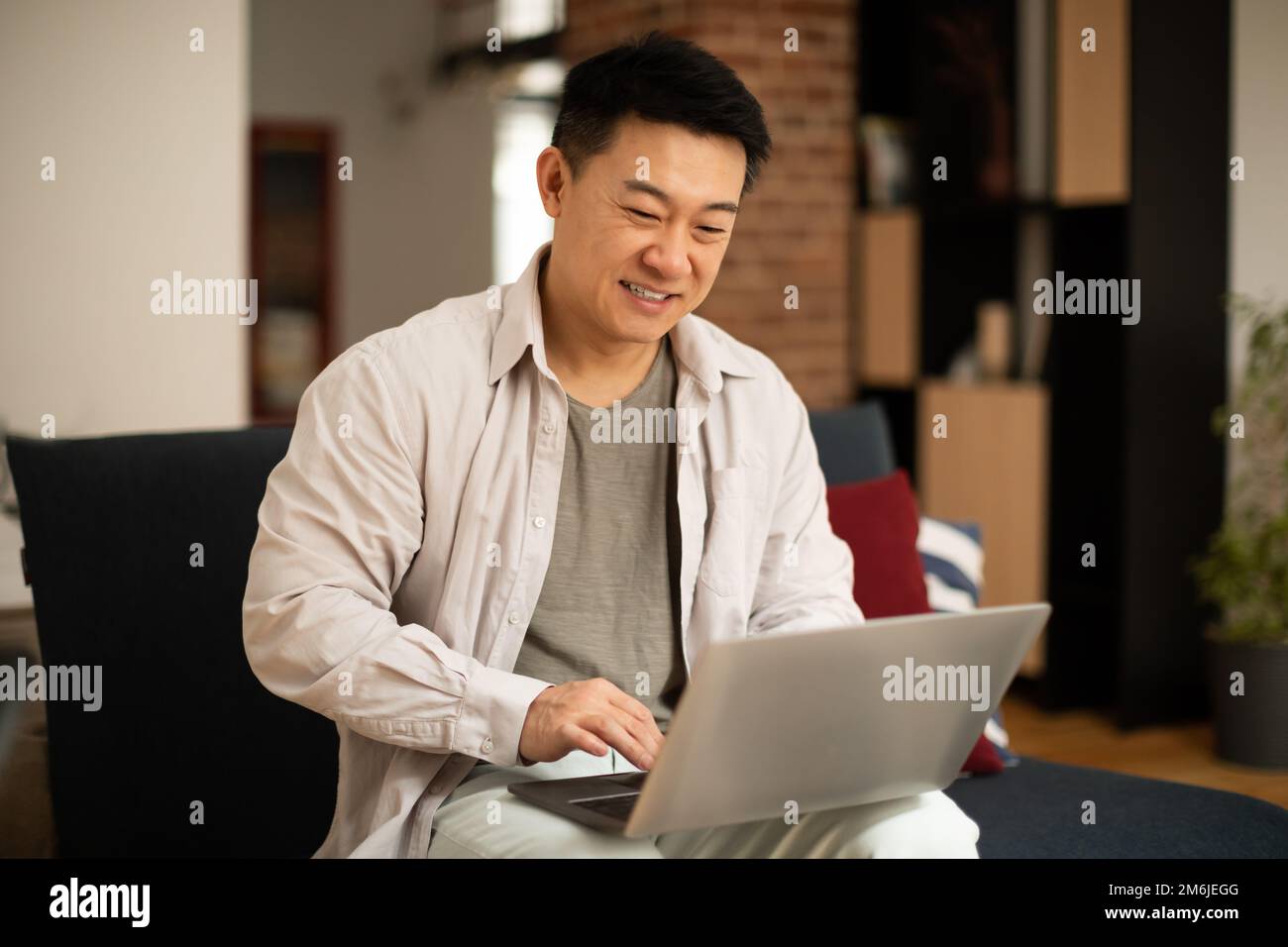 Happy asian middle aged man working on laptop, sitting on sofa in living room interior, free space Stock Photo
