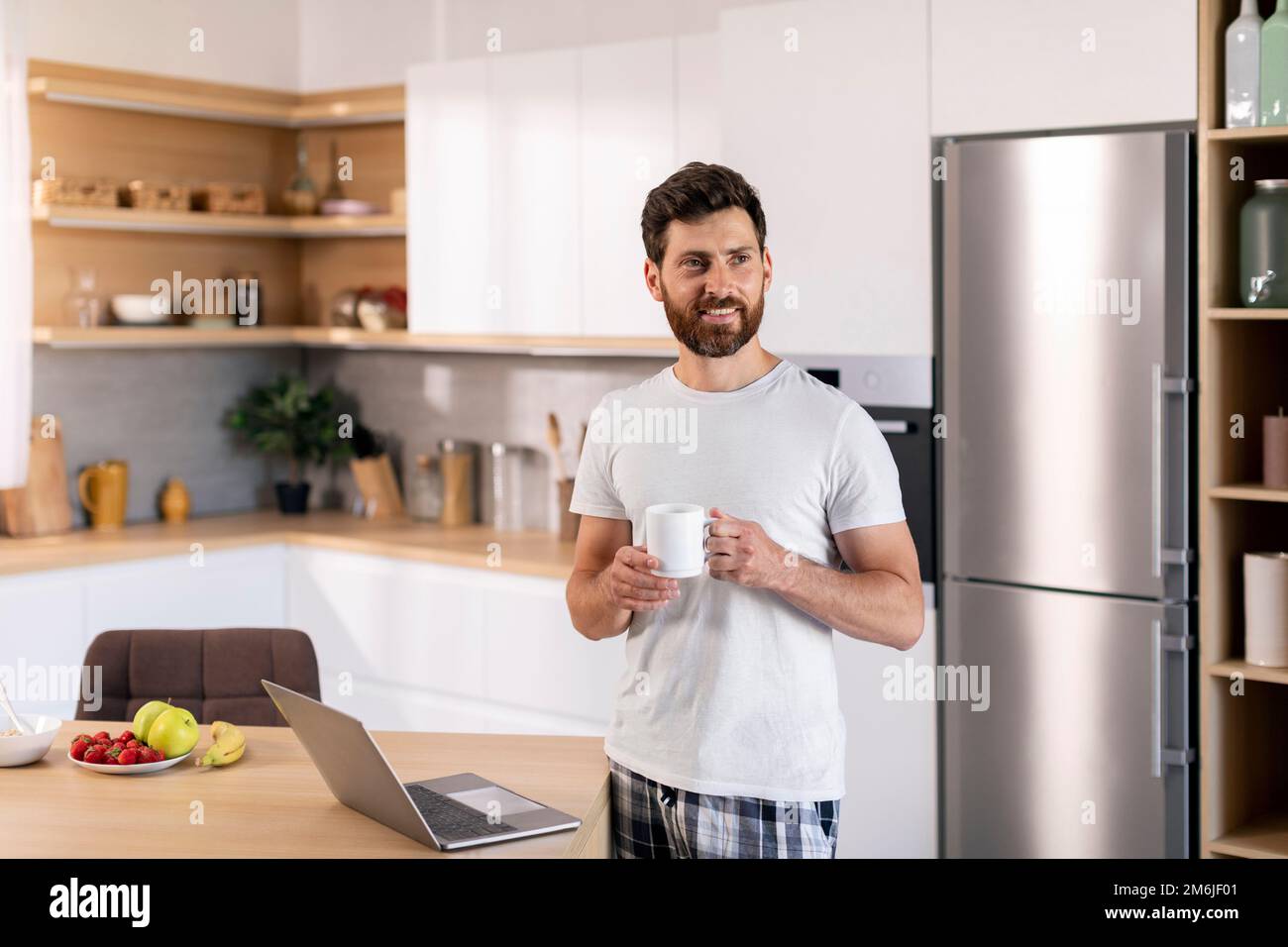 Happy millennial caucasian guy with beard with cup of coffee enjoys good morning, has breakfast with computer Stock Photo