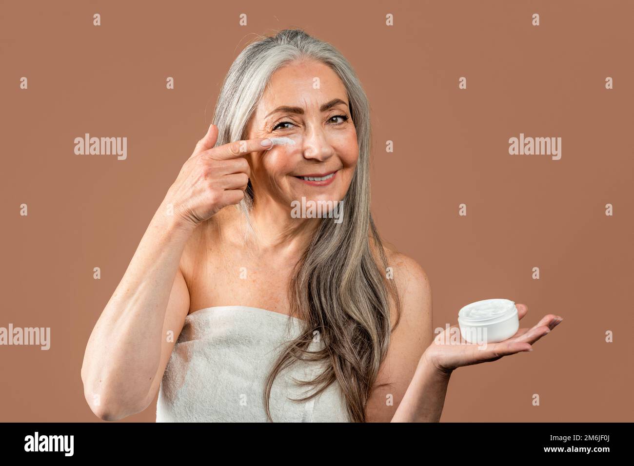 Smiling mature caucasian woman with natural beauty shows jar and applies cream on face, recommend rejuvenation Stock Photo