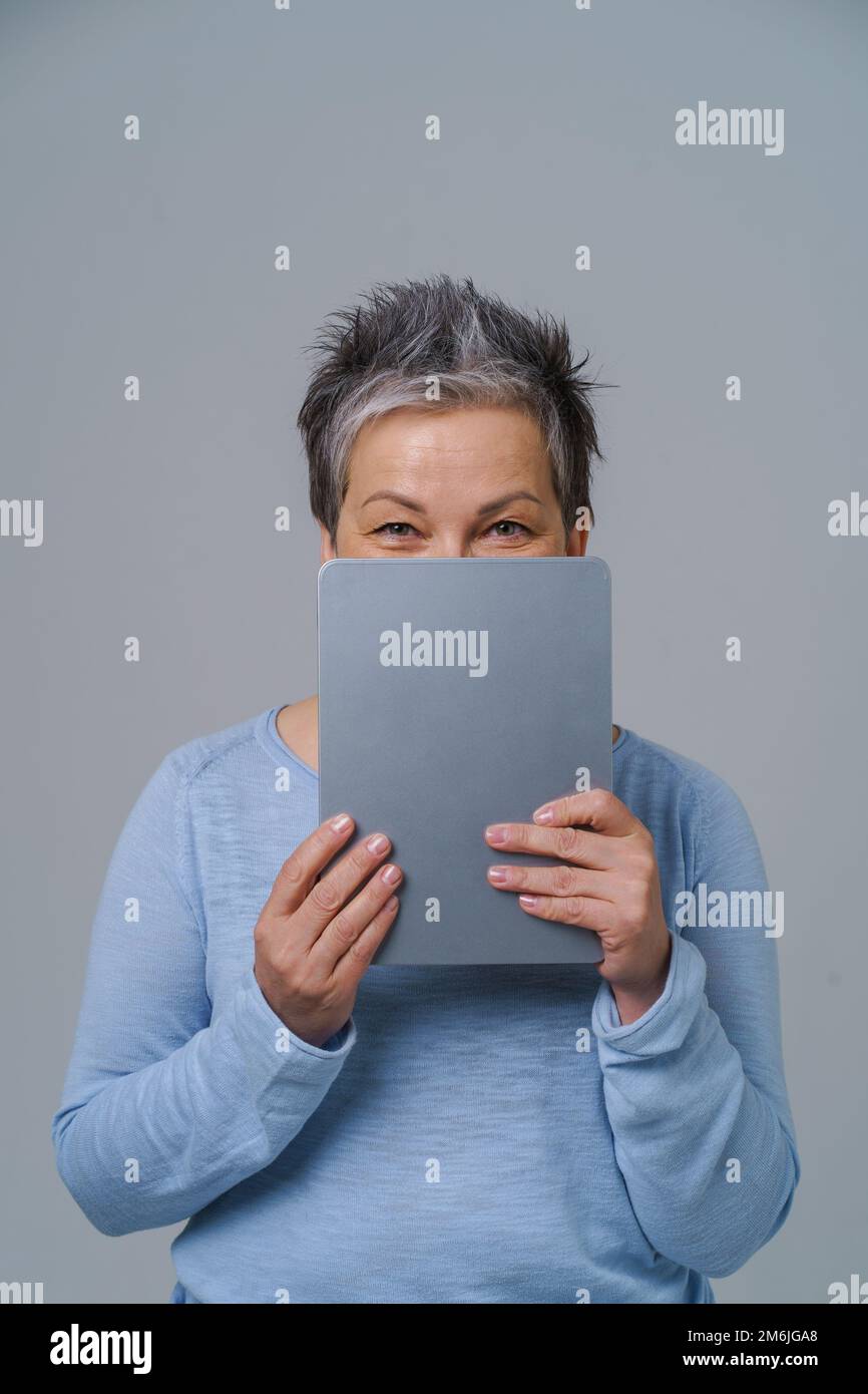Charming mature grey haired woman hide shy behind digital tablet working or shopping online or checking on social media. Pretty Stock Photo