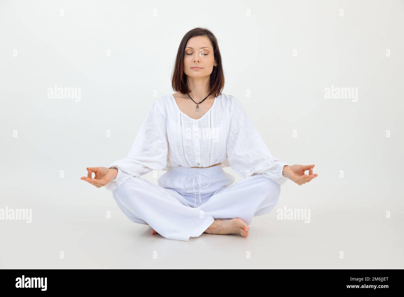 Young woman sitting with crossed legs on floor, doing Sukhasana exercise with closed eyes on white background. Yoga. Stock Photo