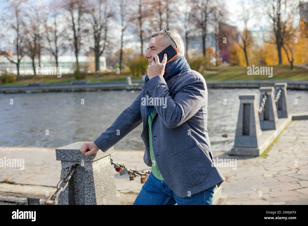 Side view of middle-aged man leaning on concrete pillars joined with chains at river in autumn, talking on smartphone. Stock Photo