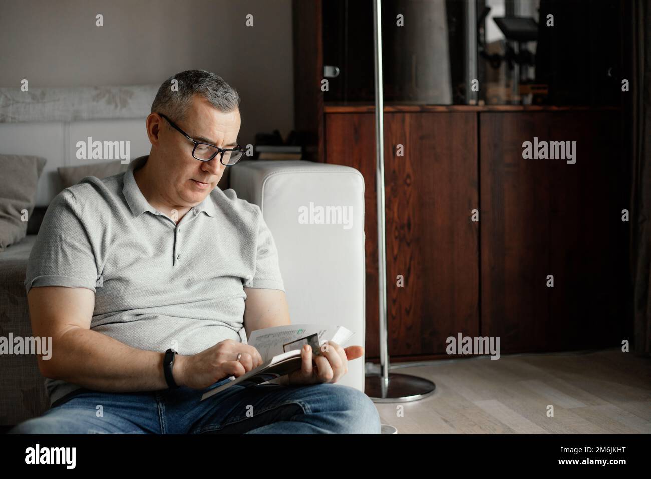 Portrait of focused middle-aged man sitting near sofa on floor, holding reading book at home. Spending free time, hobby. Stock Photo