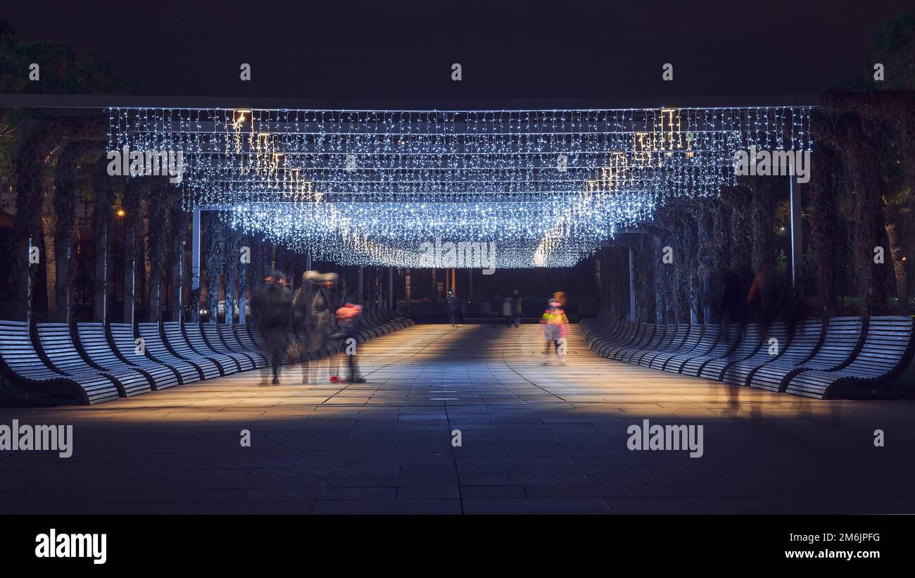 The outlines of people walking along the night alley in the park illuminated by bright hanging garlands. Selective focus. Stock Photo