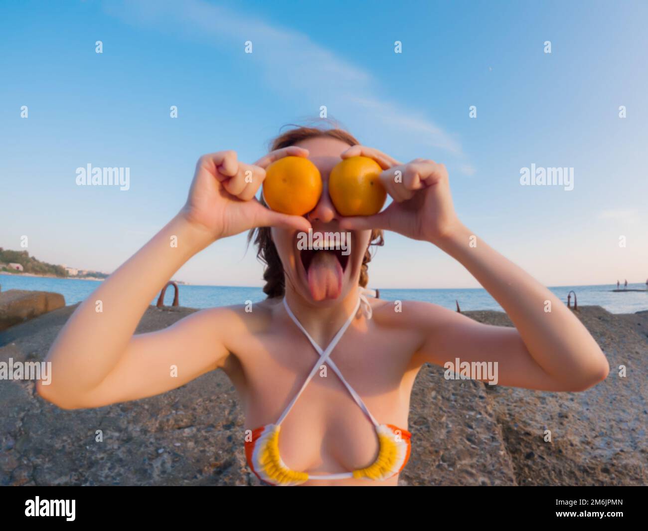 A woman shows her tongue putting tangerines to her eyes sitting on the breakwaters of the sea coast on a summer day. Humorous ph Stock Photo
