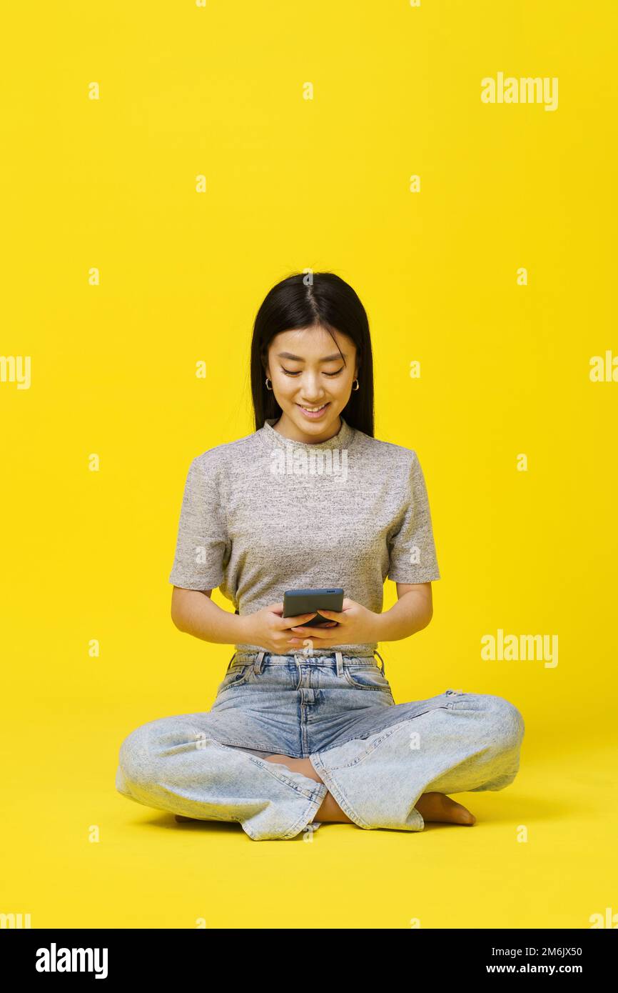 Asian young girl sitting on the floor holding smartphone texting or shopping online isolated on yellow background. Product place Stock Photo